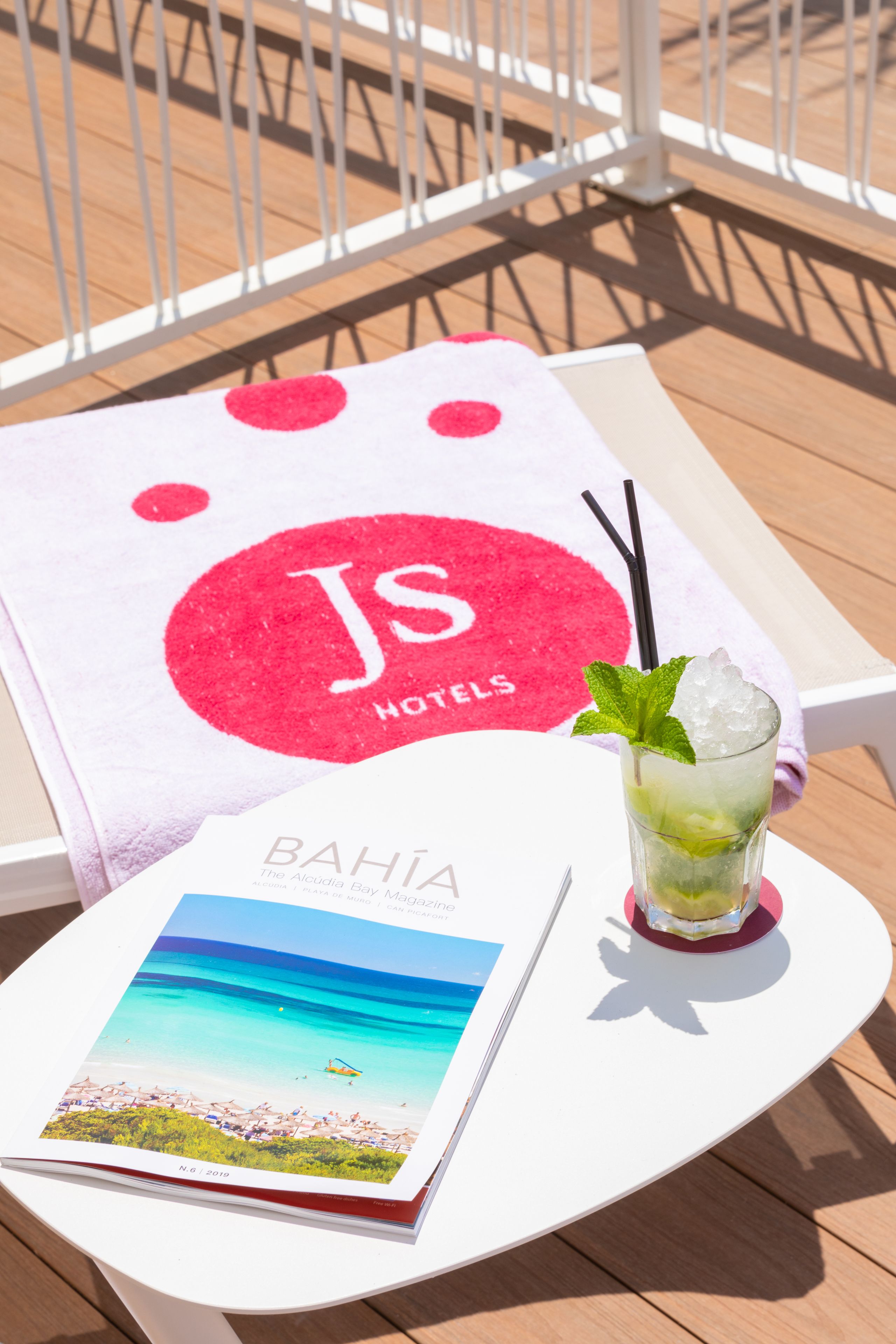 A white table with a magazine, a glass of iced drink with mint, and a towel with a logo.