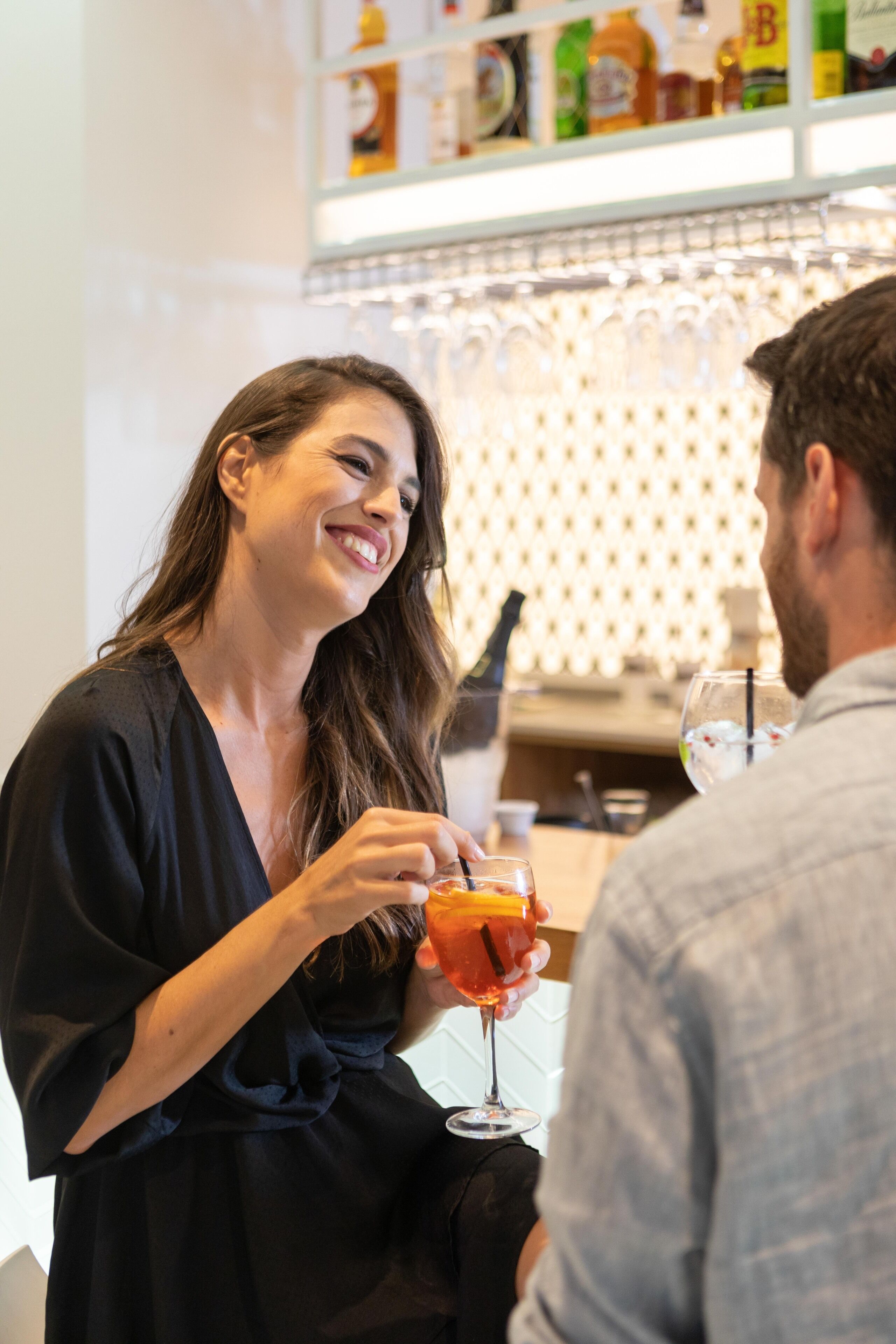 A woman holding a glass of orange drink at a bar with a shelf of bottles in the background.