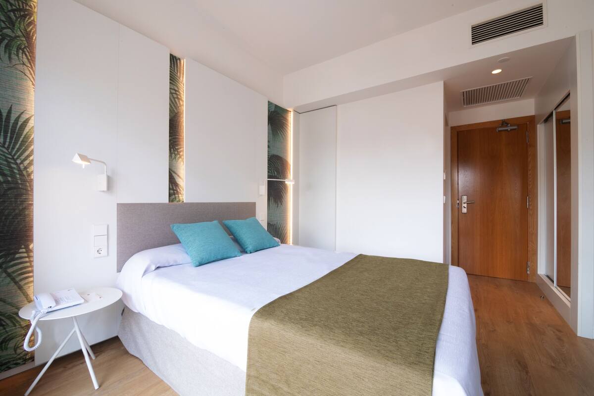 Une chambre d’hôtel avec un lit, une table de chevet avec un téléphone, une porte et un mur orné d’un motif de feuilles.