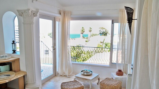 A room with a view of the beach, a small table with a tray, two wicker ottomans, and a white column. - Uma Casa à Beira Sol (Lagos)