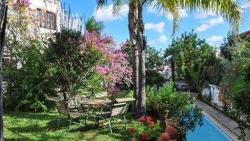Zona junto a la piscina con mesa y sillas de madera, rodeada de abundante follaje y plantas en flor.