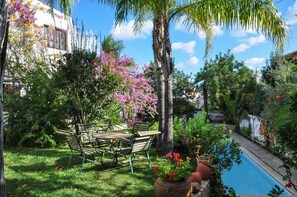 Zona junto a la piscina con mesa y sillas de madera, rodeada de abundante follaje y plantas en flor.