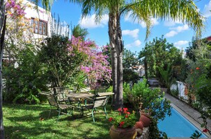 A poolside area with a wooden table and chairs, surrounded by lush greenery and flowering plants.