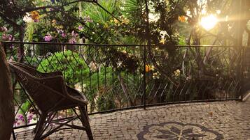 Una silla de mimbre en un patio con baldosas, una cerca de metal y abundante follaje al fondo.