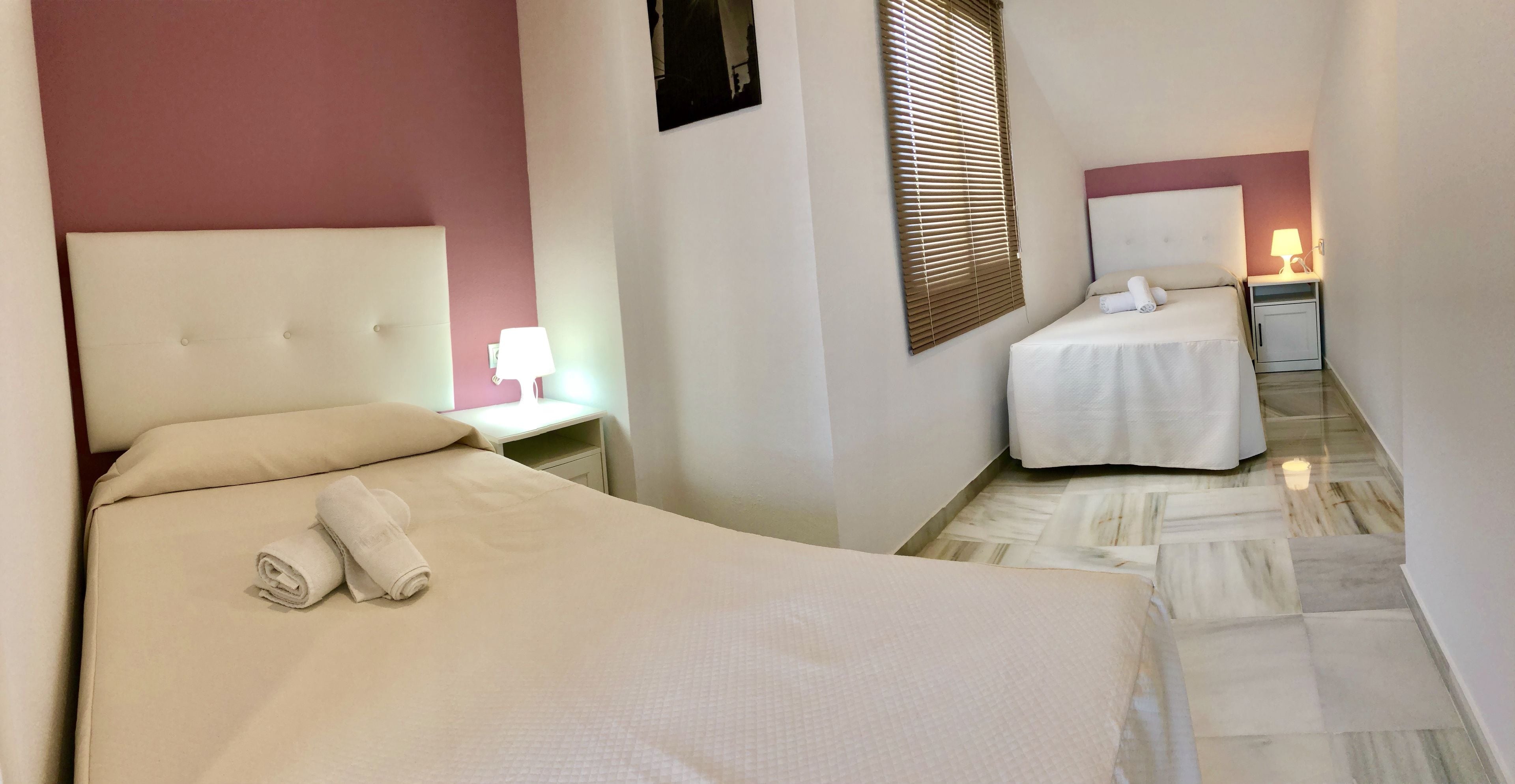 Ein Hotelzimmer mit einem großen Bett, einem kleineren Bett, einem Nachttisch mit Lampe und einem Fenster mit Jalousien.