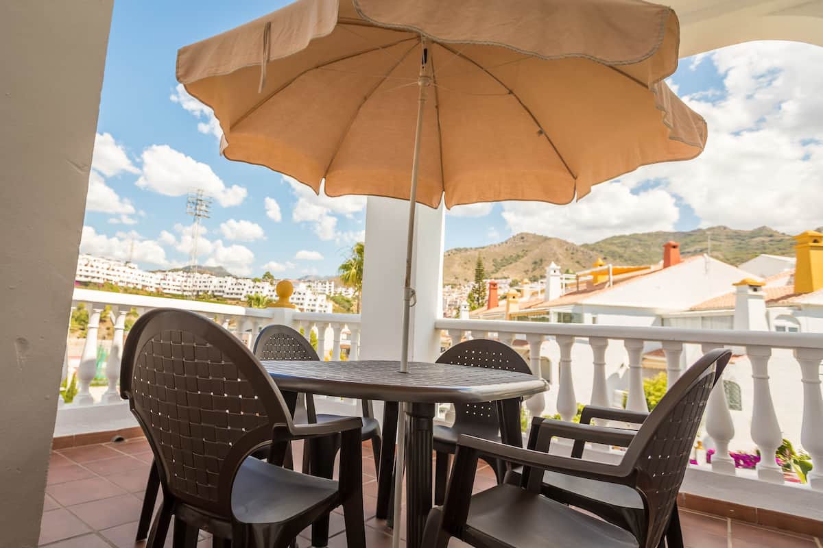 Een terras met een tafel, stoelen, een parasol en uitzicht op gebouwen en bergen.