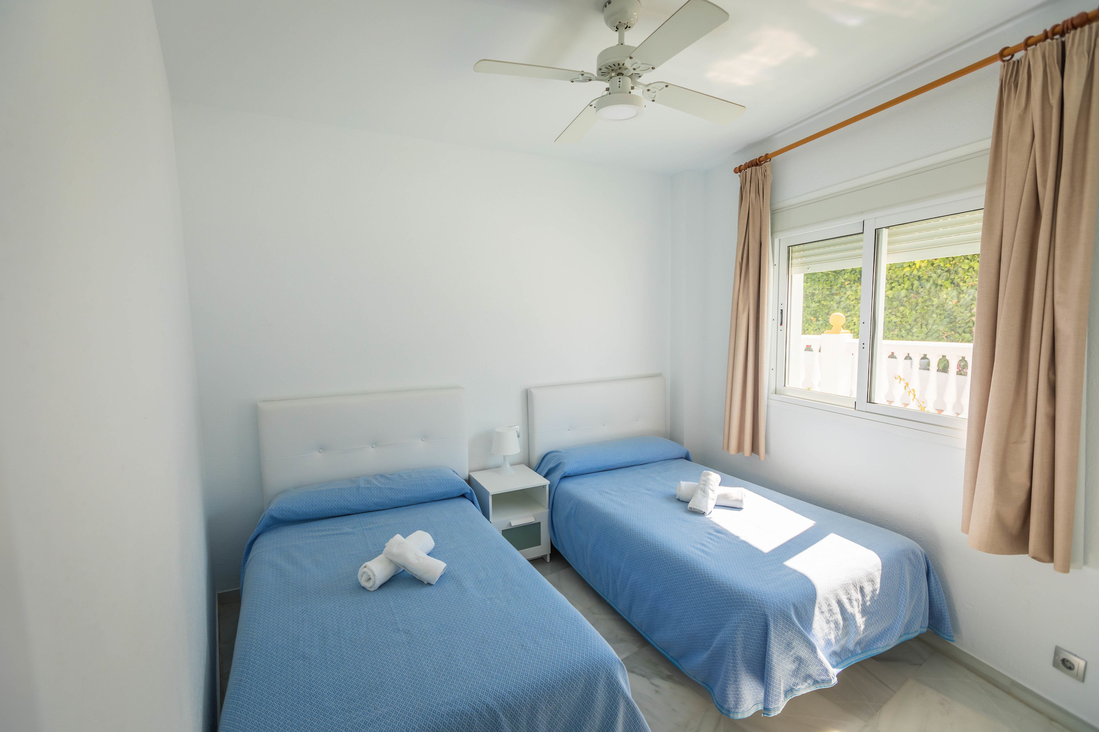 Ein Schlafzimmer mit zwei Einzelbetten, einem Deckenventilator, einem Nachttisch und einem Fenster mit Vorhängen.