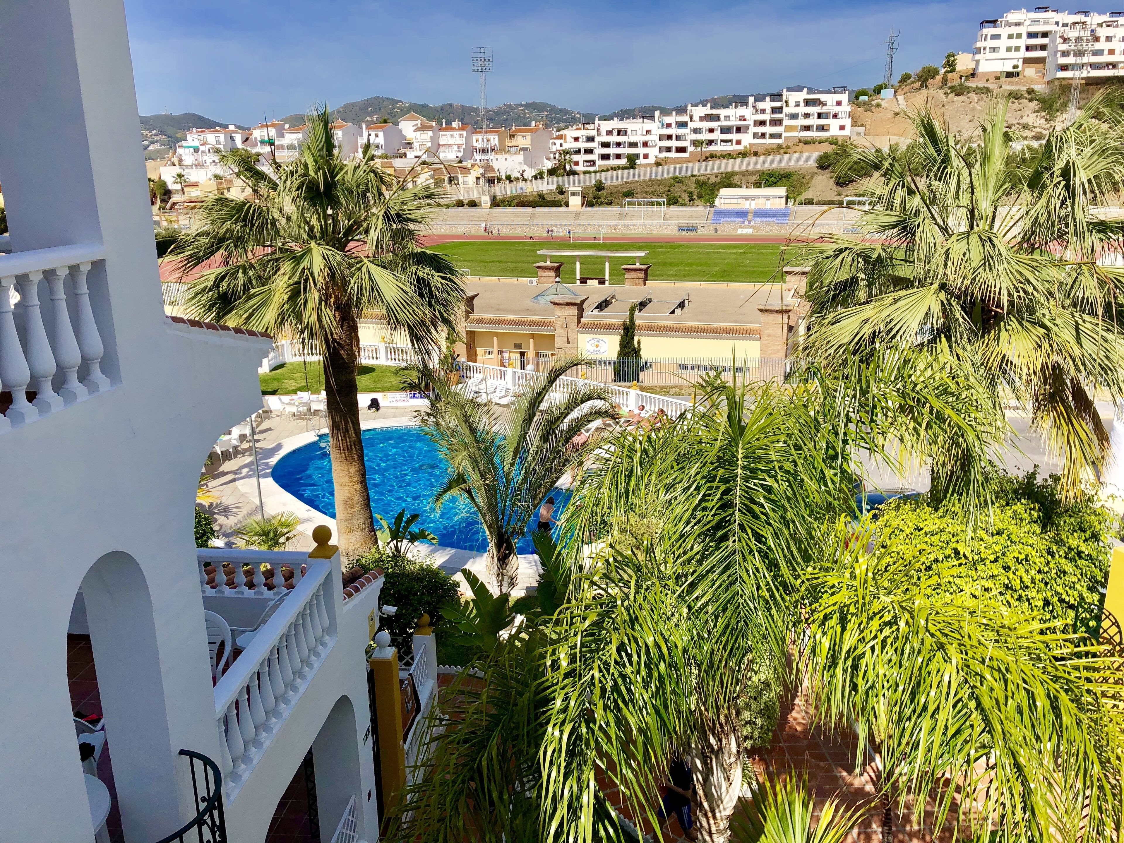 Zona de piscina con palmeras, cancha de tenis y vistas a un área residencial con edificios.