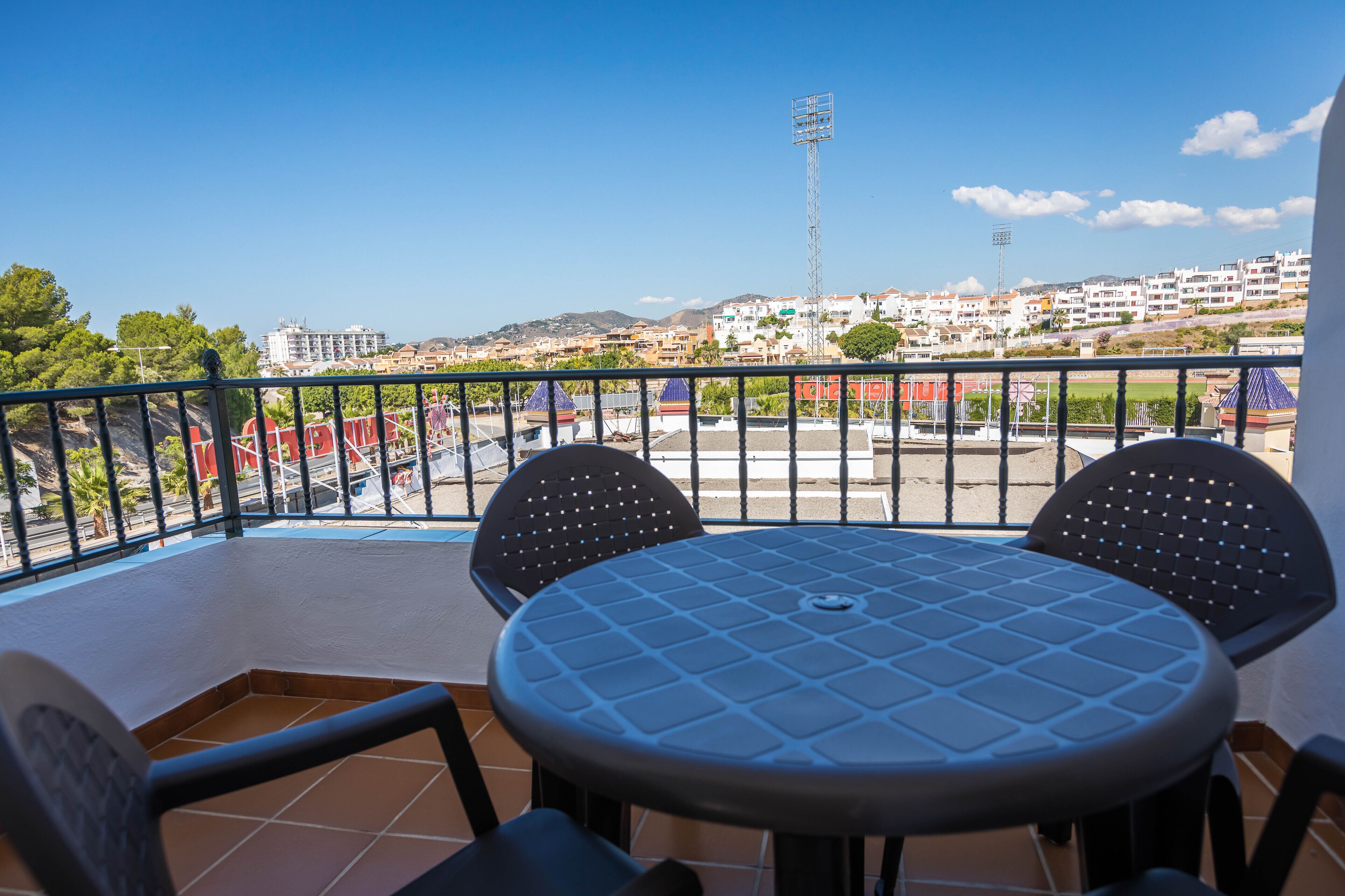 Un balcon avec une table ronde et deux chaises, donnant sur un quartier résidentiel sous un ciel dégagé.