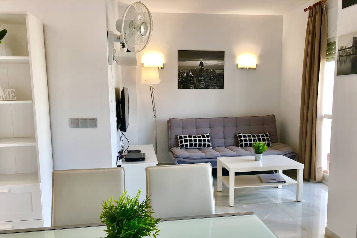 Een moderne woonkamer met een grijze bank, een witte salontafel, een eettafel met glazen blad, een potplant, een ventilator aan de muur, een flatscreen televisie en twee wandlampen.