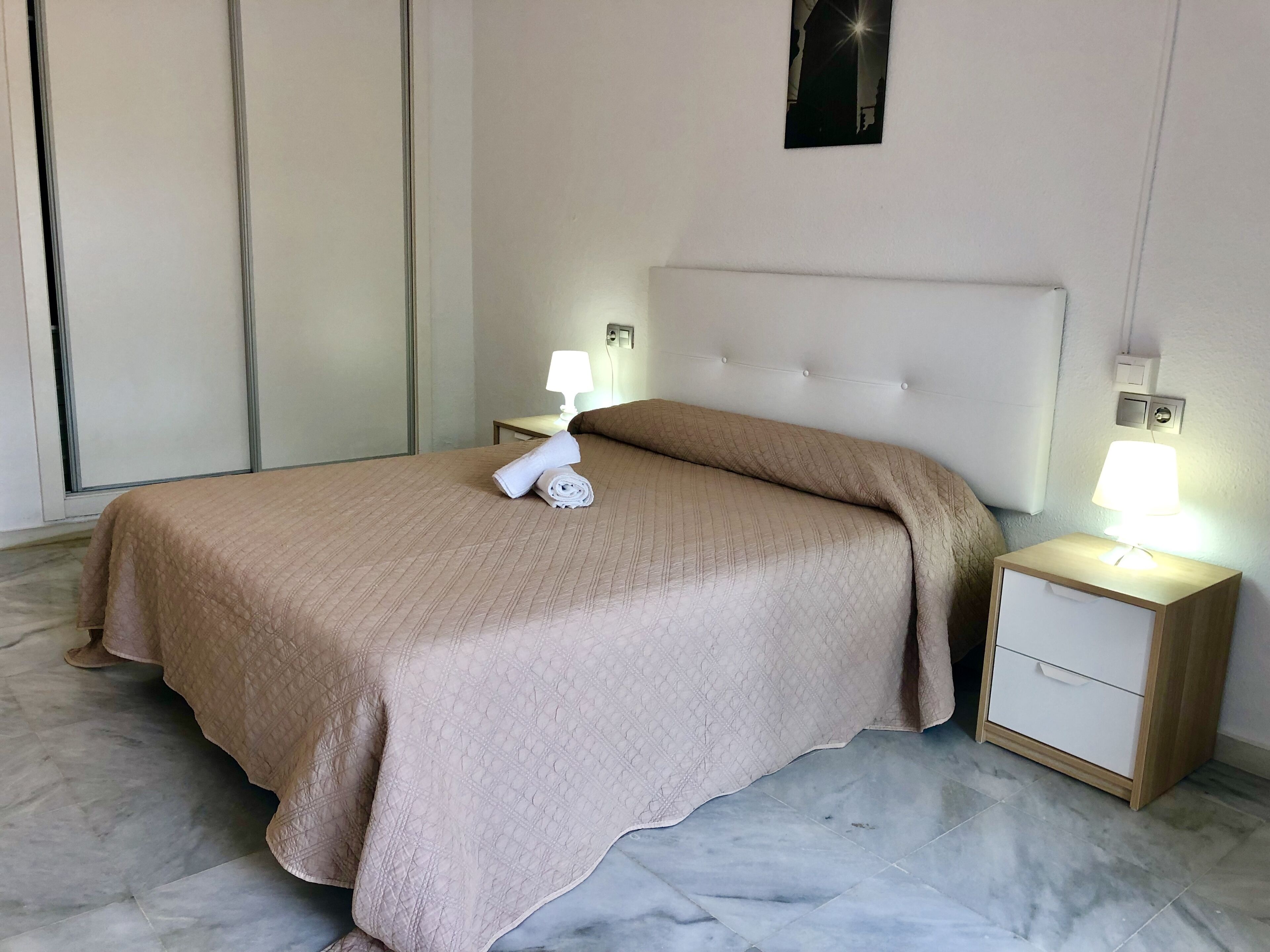 A neatly made bed with a beige quilt, two rolled towels, and two bedside lamps on wooden nightstands.