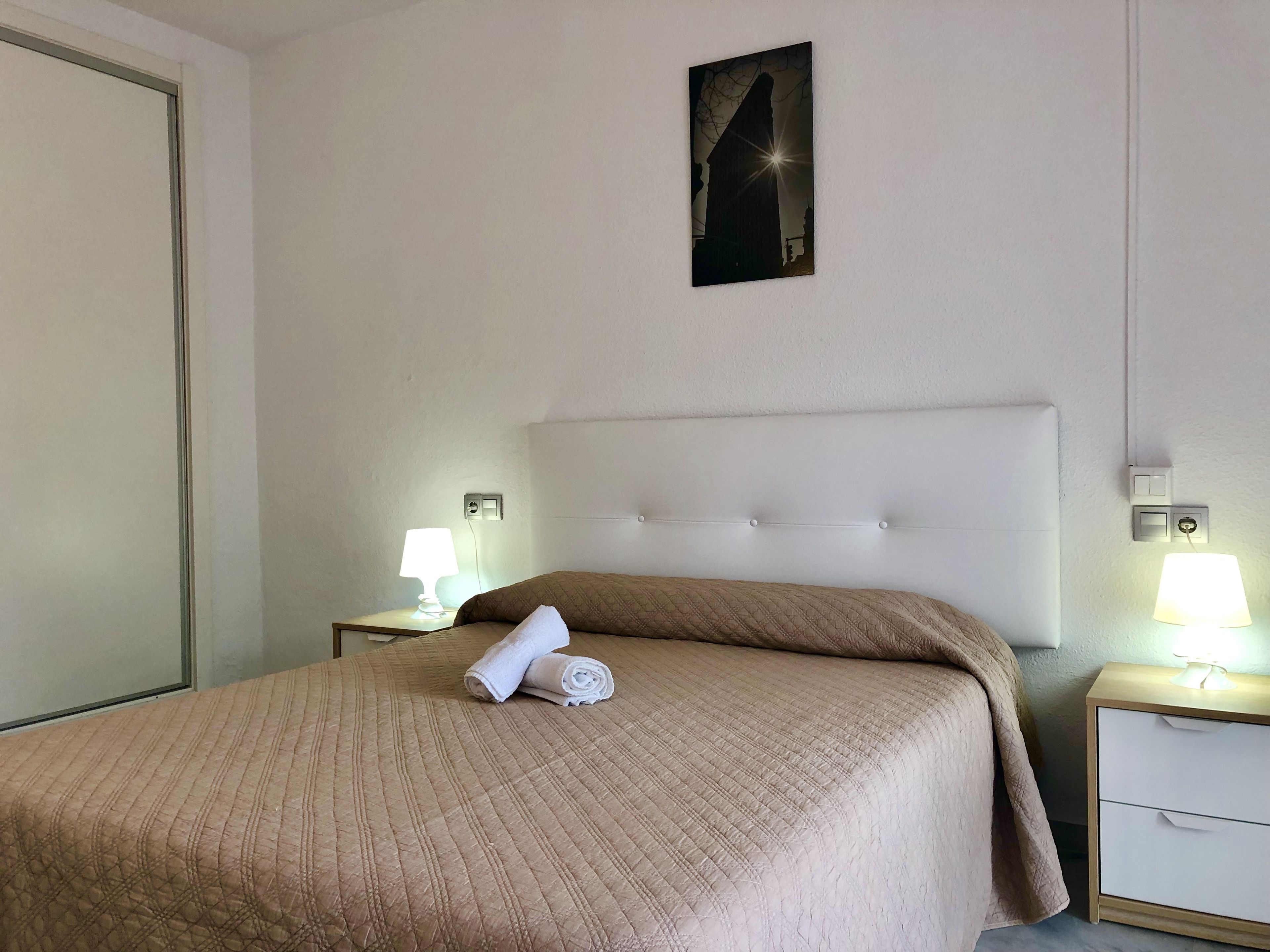 Habitación de hotel con cama, dos mesitas de noche con lámparas, un cuadro colgado en la pared y una puerta.