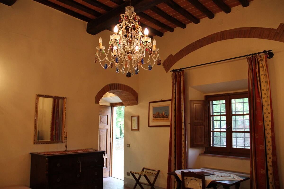 A room with a chandelier, wooden beams, a dresser, a chair, a table, a window with curtains, and a door leading outside.