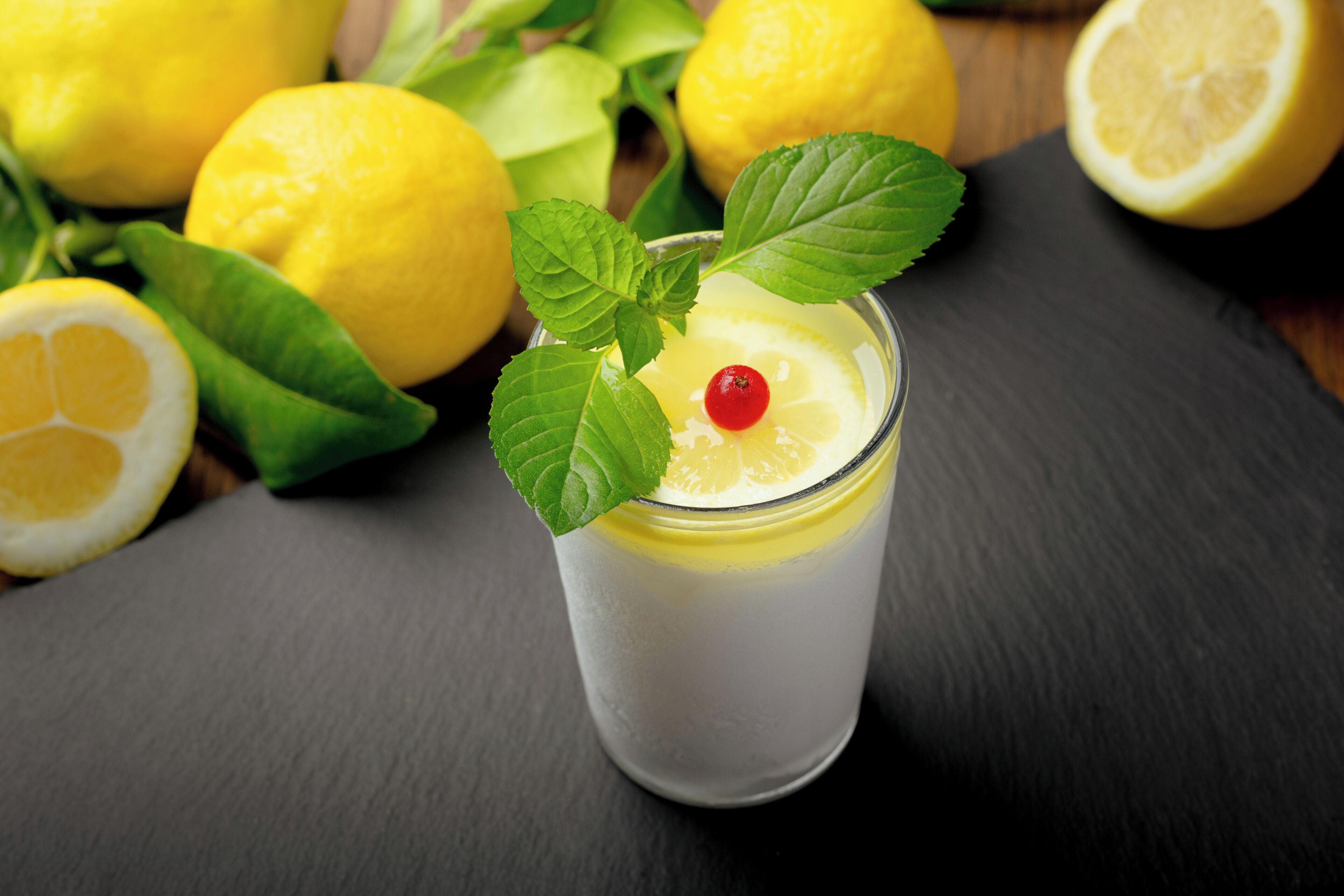 A glass of a clear beverage with a slice of lemon and a cherry on top, garnished with mint leaves, on a dark surface with lemons and lemon slices around.