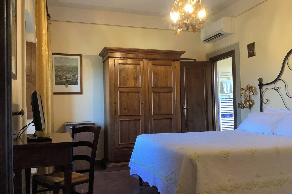 A bedroom with a bed, a wooden wardrobe, a desk with a chair, and a chandelier.
