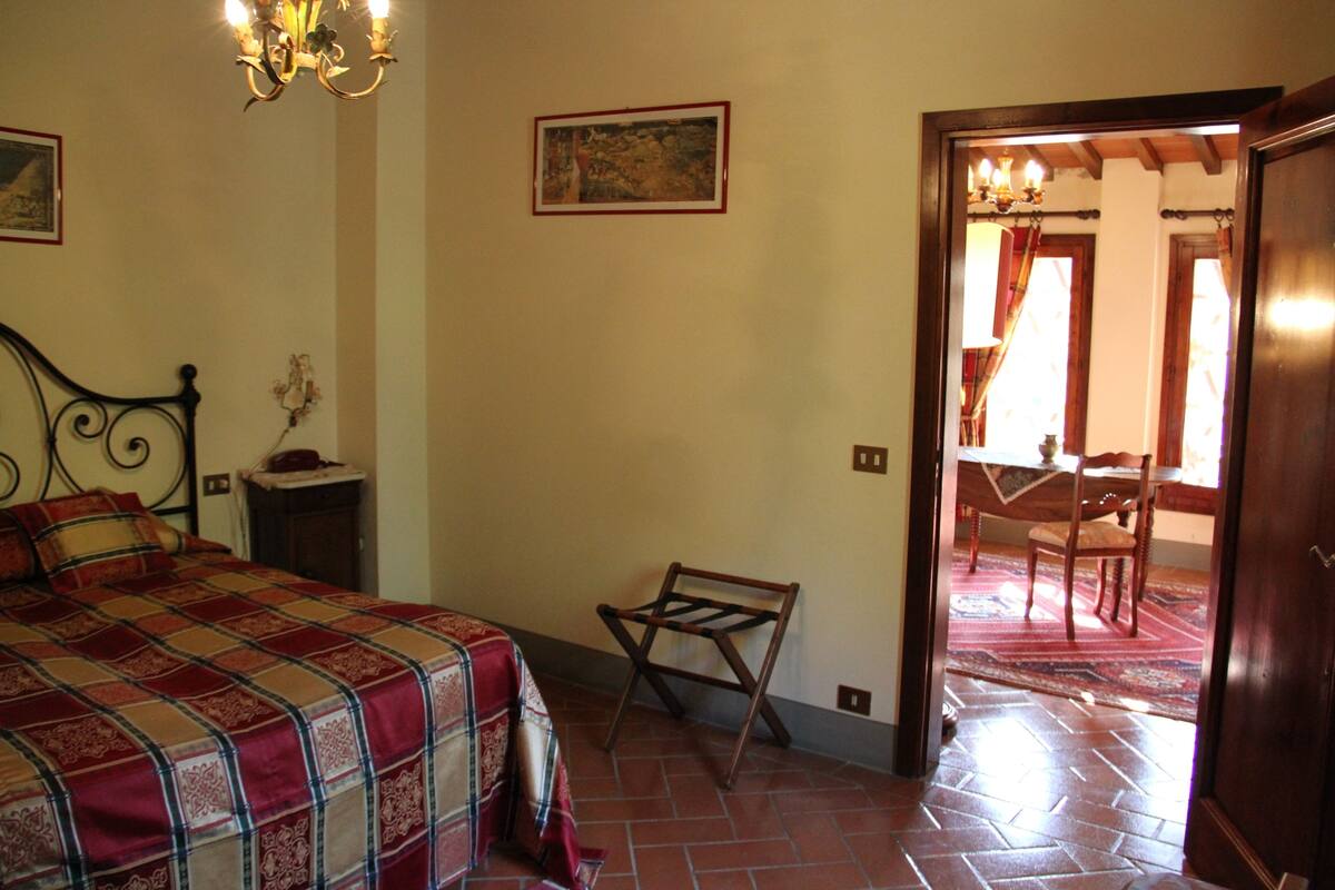 A bedroom with a bed, a nightstand, a chair, a wooden door, and a view of a dining area.