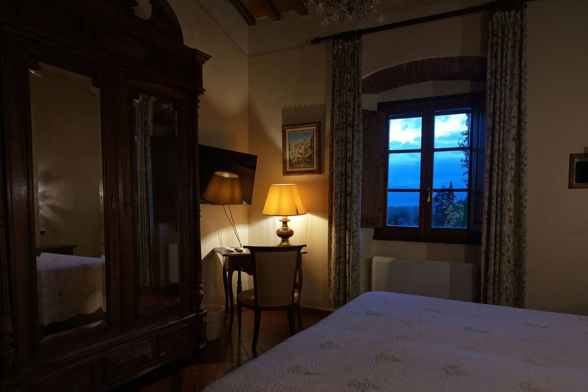 A hotel room with a bed, a desk with a lamp, a large mirror, a painting on the wall, and a window with a view.