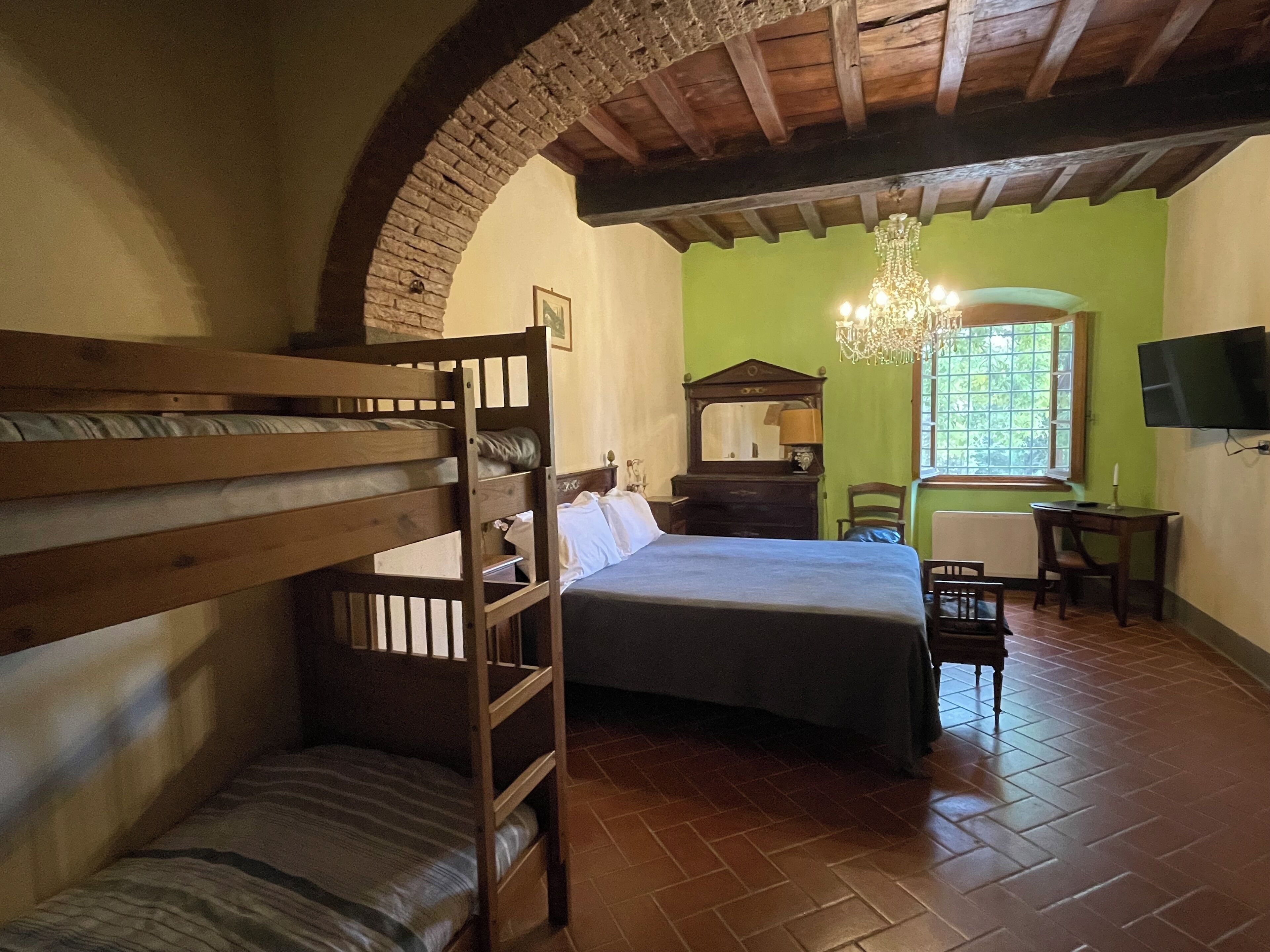 Una habitación con literas, una cama individual, un escritorio de madera, una silla, un espejo, una araña y un televisor en la pared.