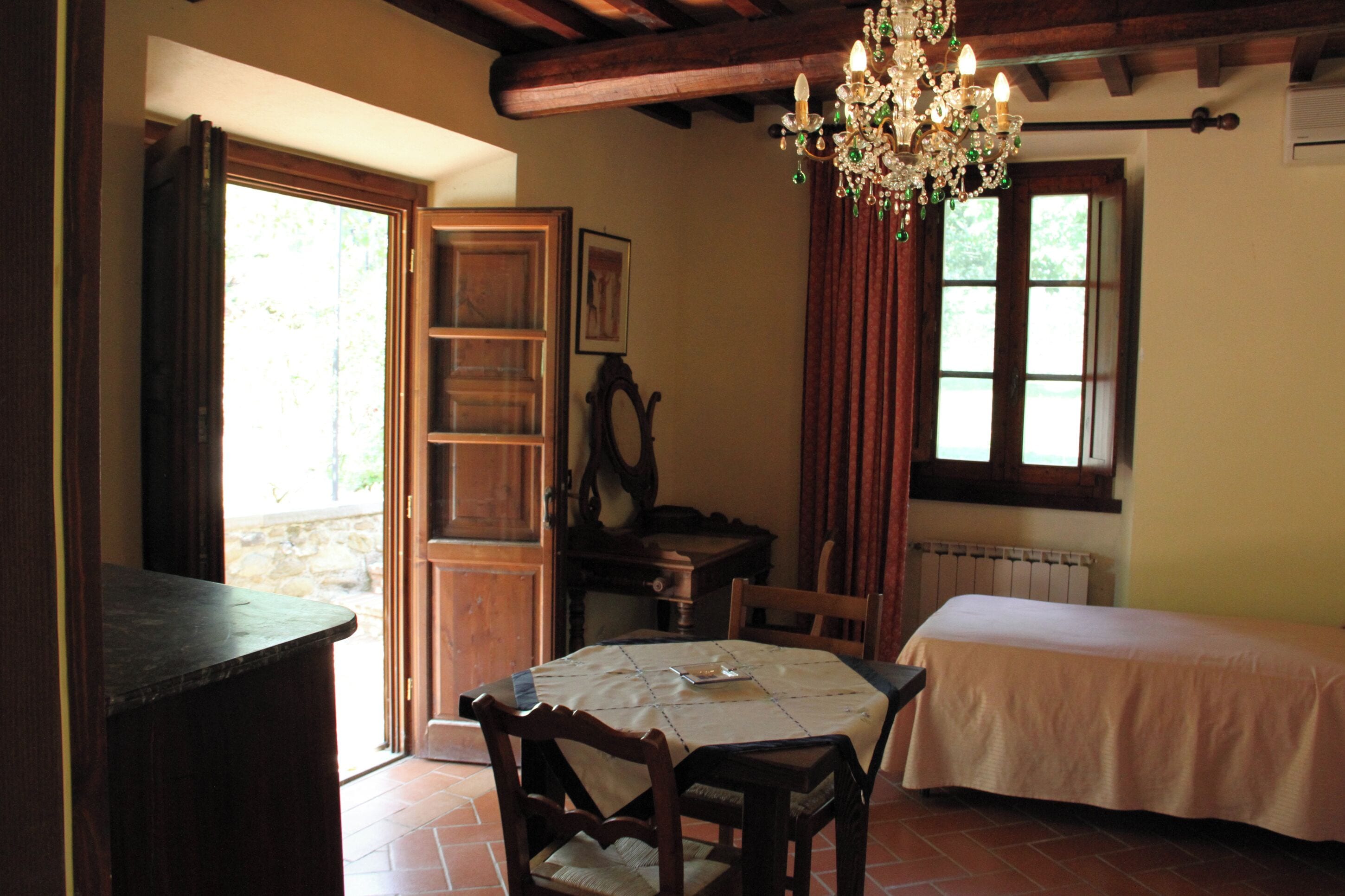 A room with a bed, a table with chairs, a wooden cabinet, a chandelier, and a window with curtains.