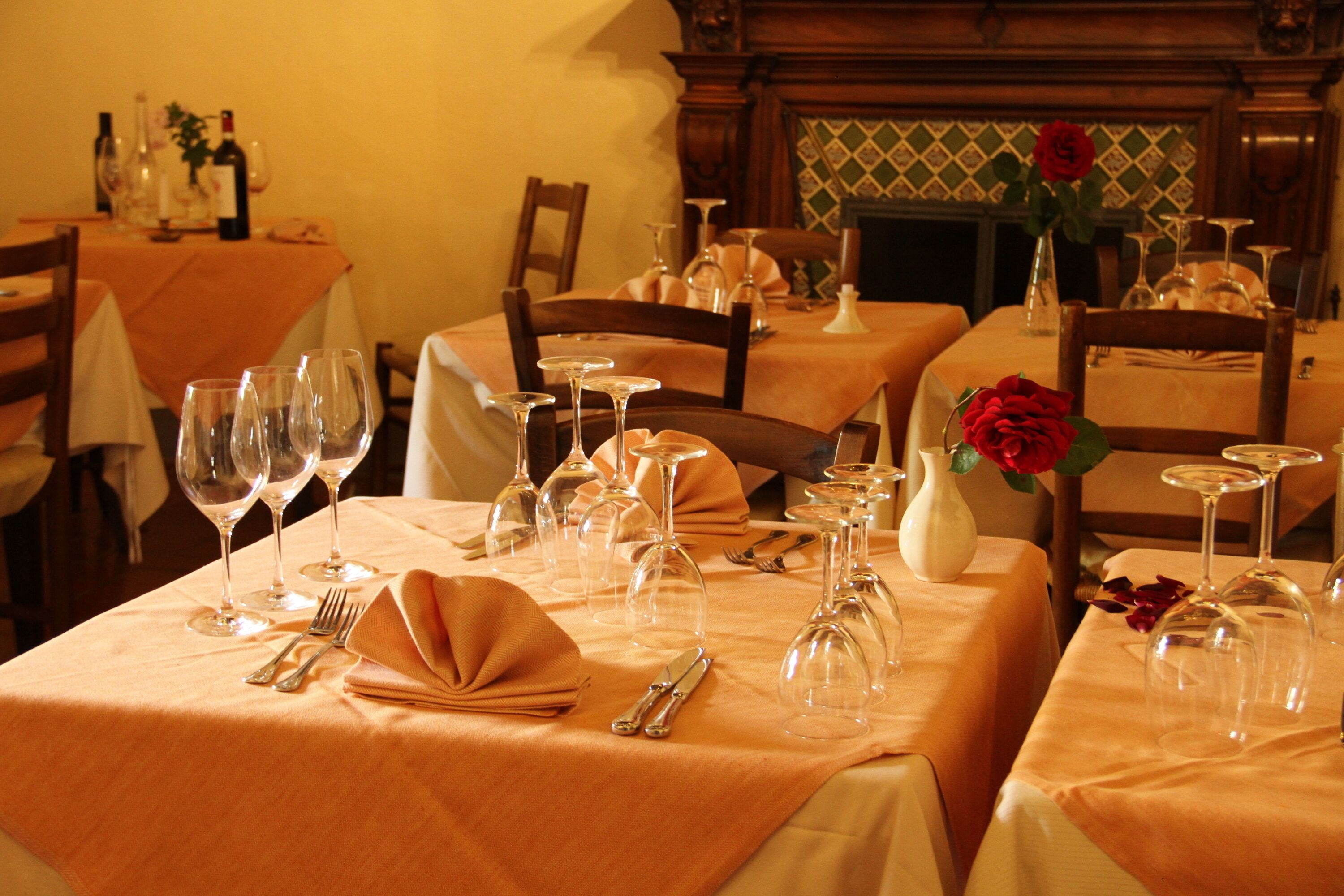 A dining area with tables set for a meal, featuring wine glasses, cutlery, and a vase with a red rose.