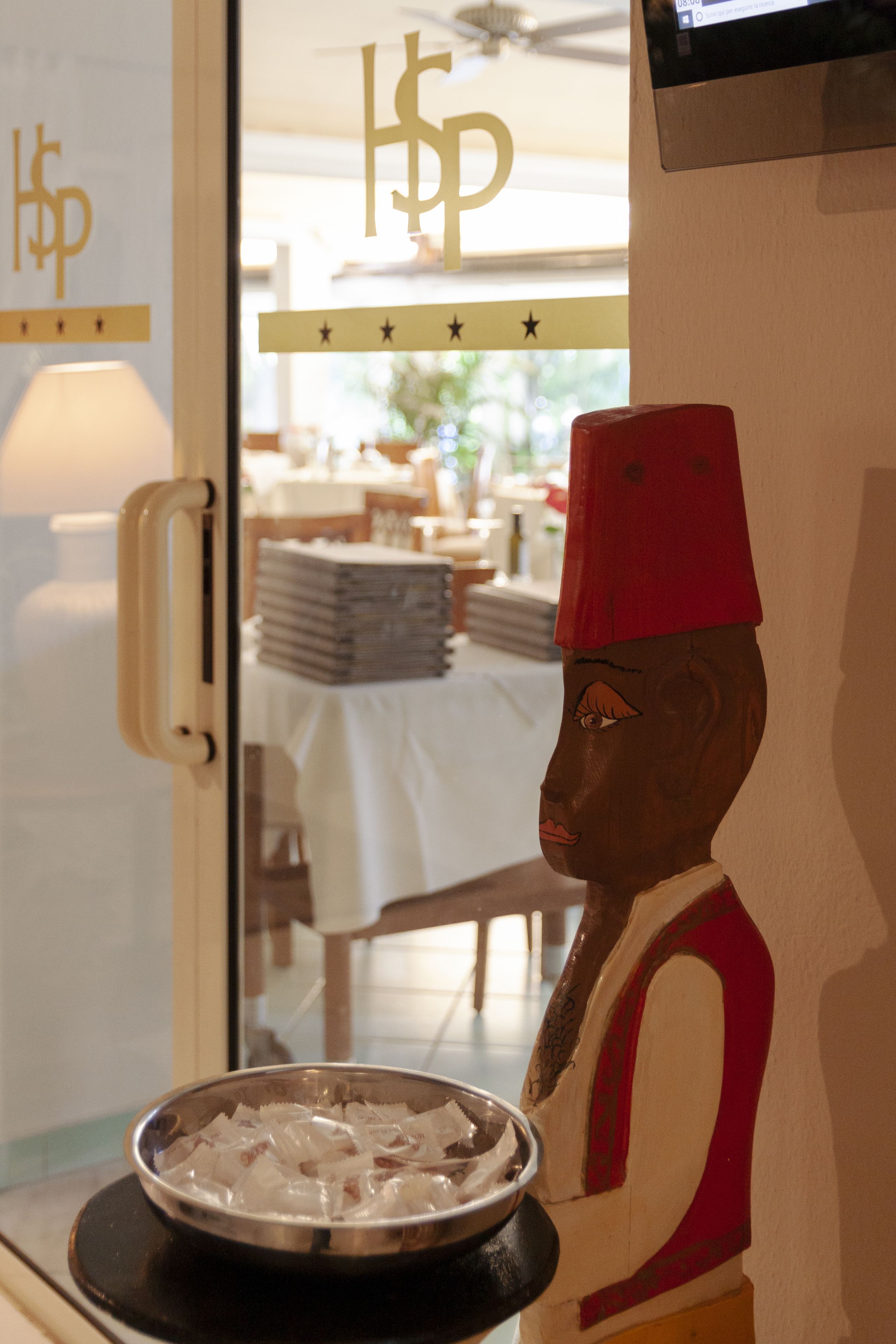 Een hotel receptie met een decoratief beeld, een metalen schaal op een balie en een tafel met gestapelde voorwerpen op de achtergrond.