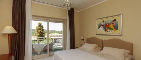A hotel room with a large bed, a chandelier, a painting on the wall, and a balcony with a view of a tree and buildings.