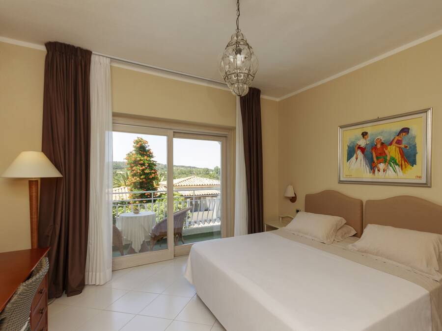 Habitación de hotel con una cama grande, un candelabro, un cuadro en la pared y un balcón con vistas a un árbol y edificios.
