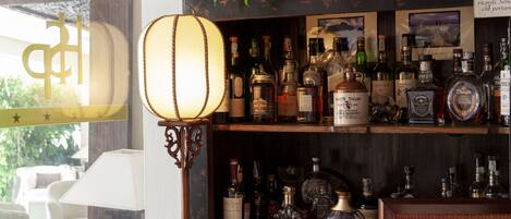 Zona de bar con estantes llenos de botellas de licor, una lámpara colgante y un letrero con texto.