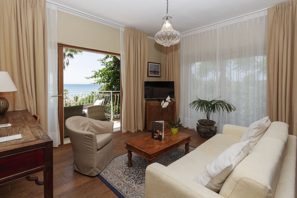 A living room with a sofa, armchair, wooden coffee table, and a TV. There is a balcony with a view of the sea.