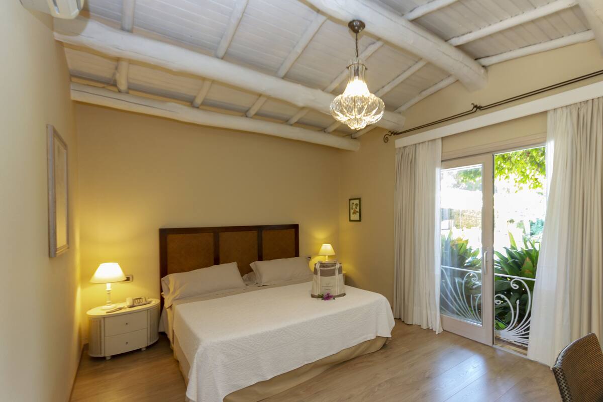 A hotel room with a bed, bedside lamps, a nightstand, a chandelier, and a view through a glass door.