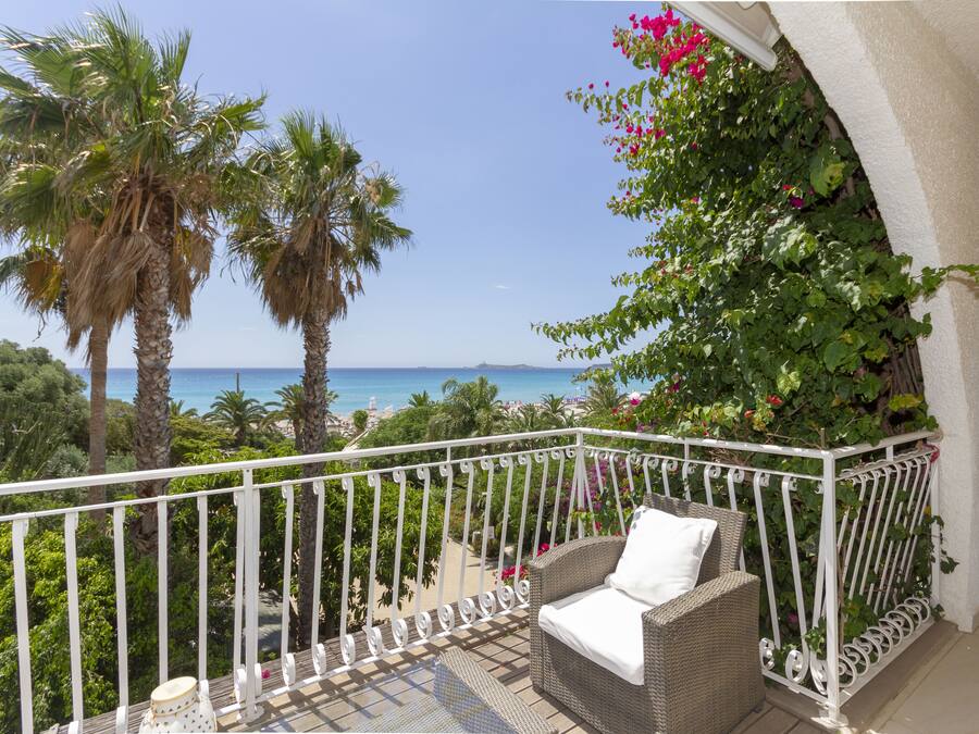 A balcony with a wicker sofa and a white cushion, overlooking a beach with palm trees and a clear blue sky.