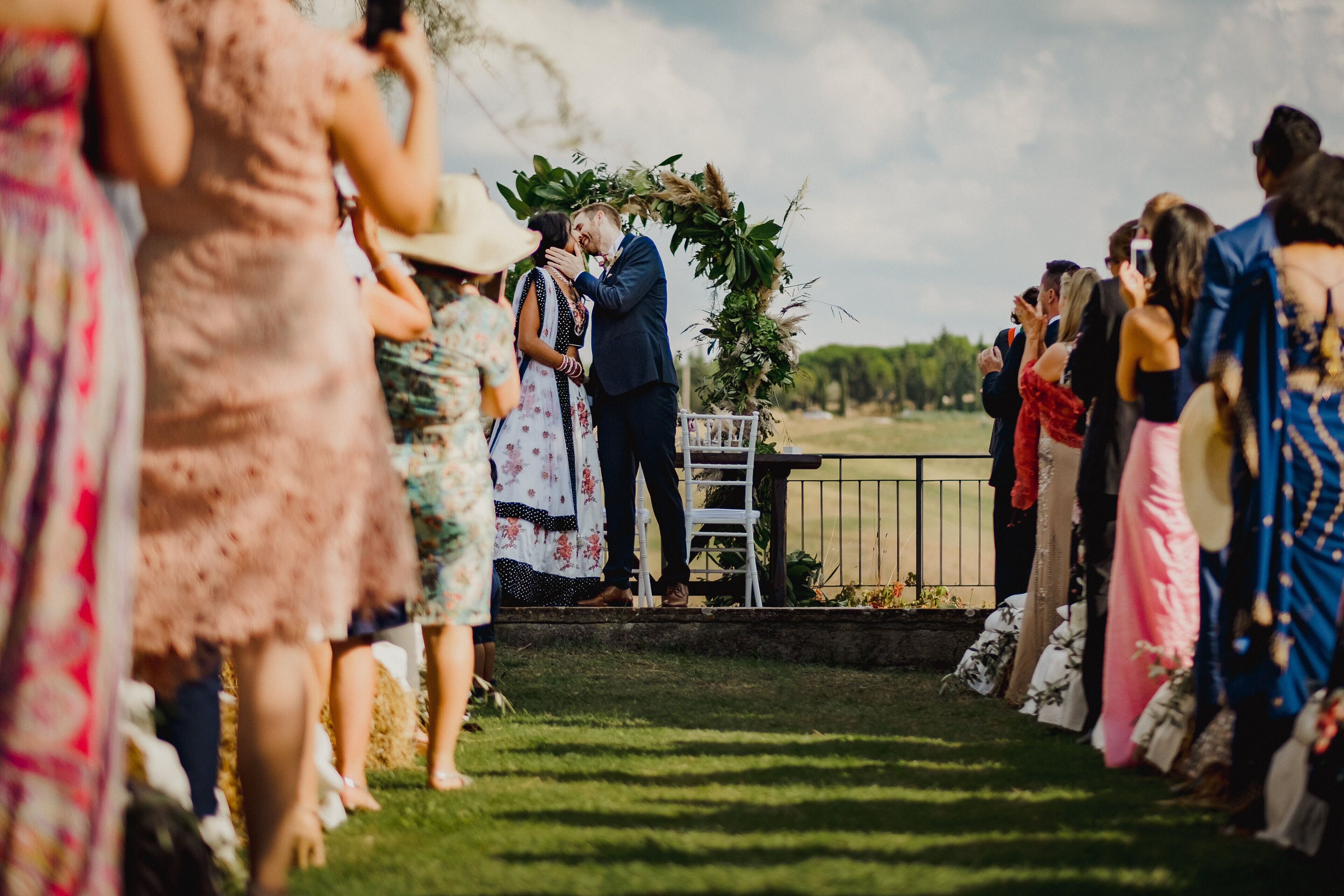 Une cérémonie de mariage où les invités sont rassemblés en demi-cercle, un couple s’embrasse et une arche décorée se trouve en arrière-plan.