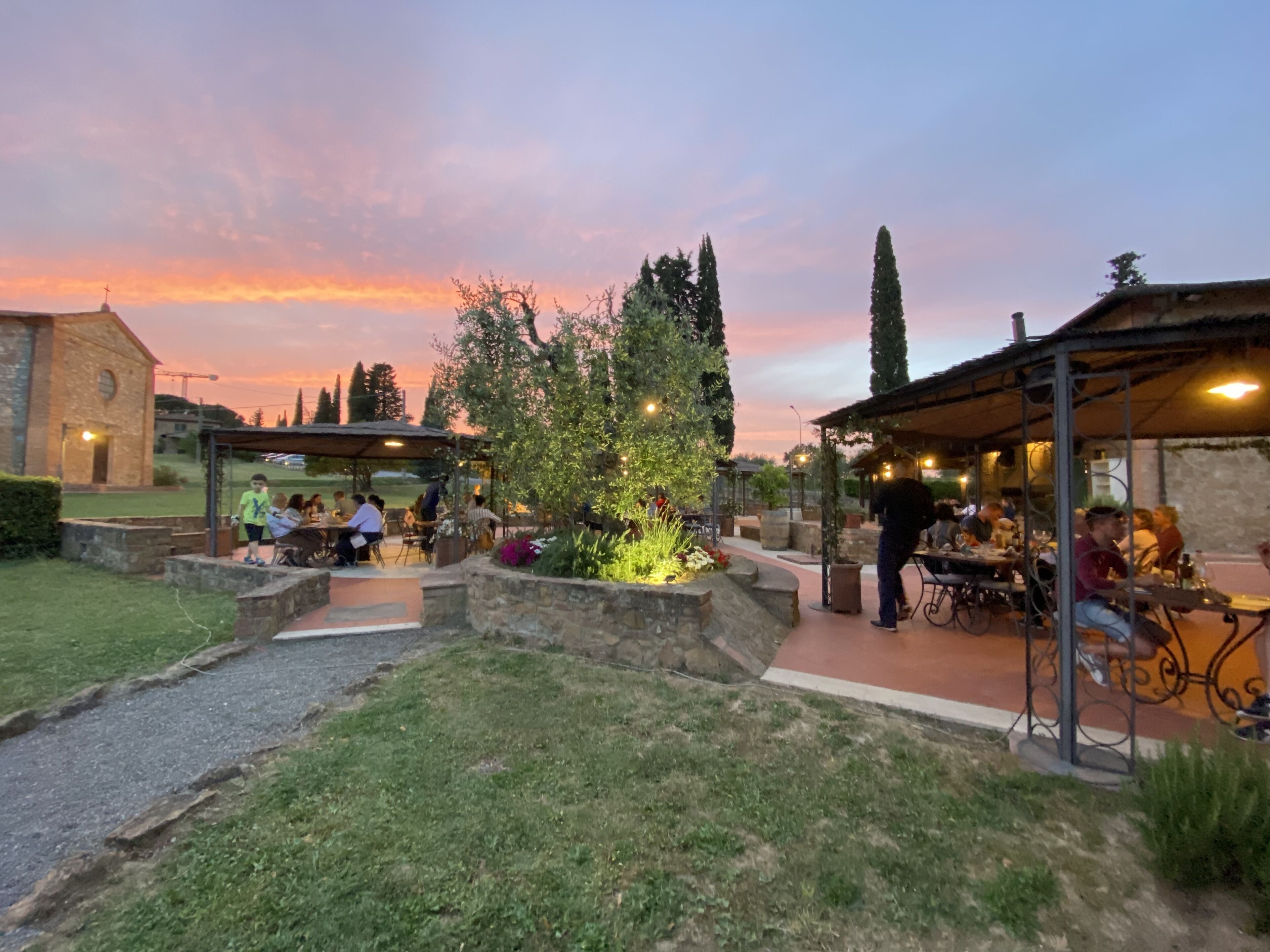 An outdoor dining area with stone walls and greenery, featuring tables and chairs under a covered structure.