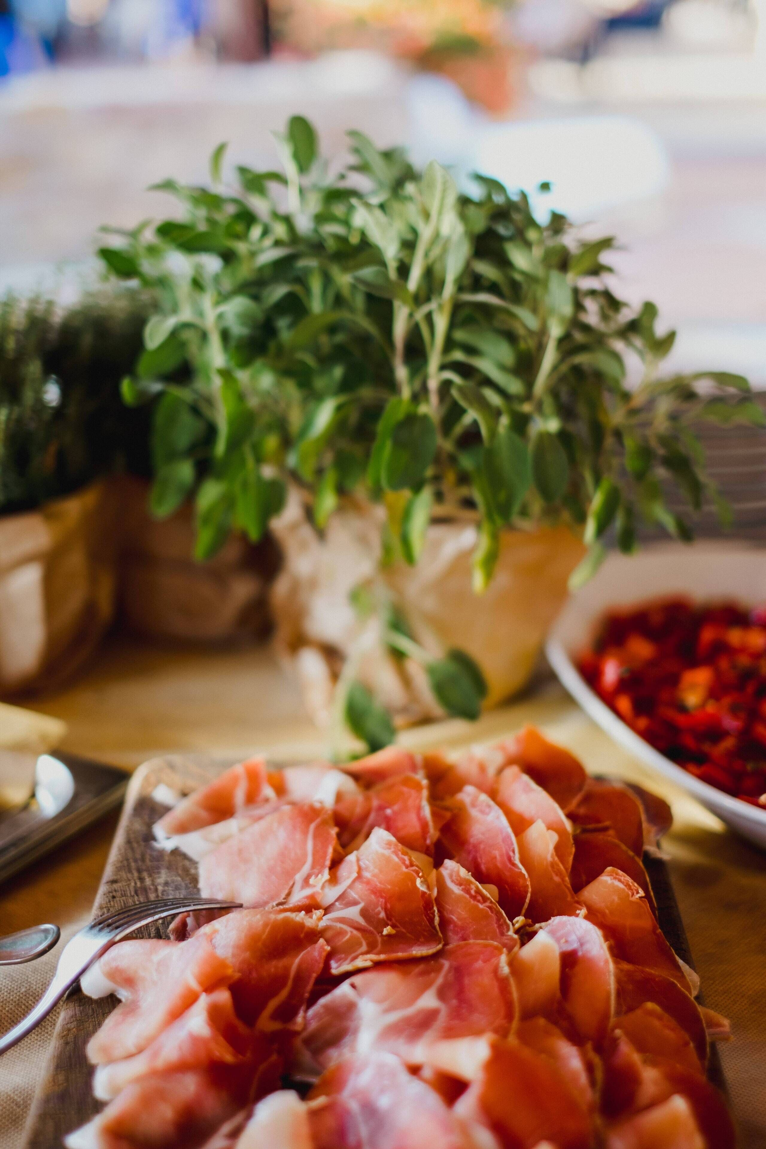 A wooden table with a platter of sliced cured meat, a bowl of red peppers, and potted plants.
