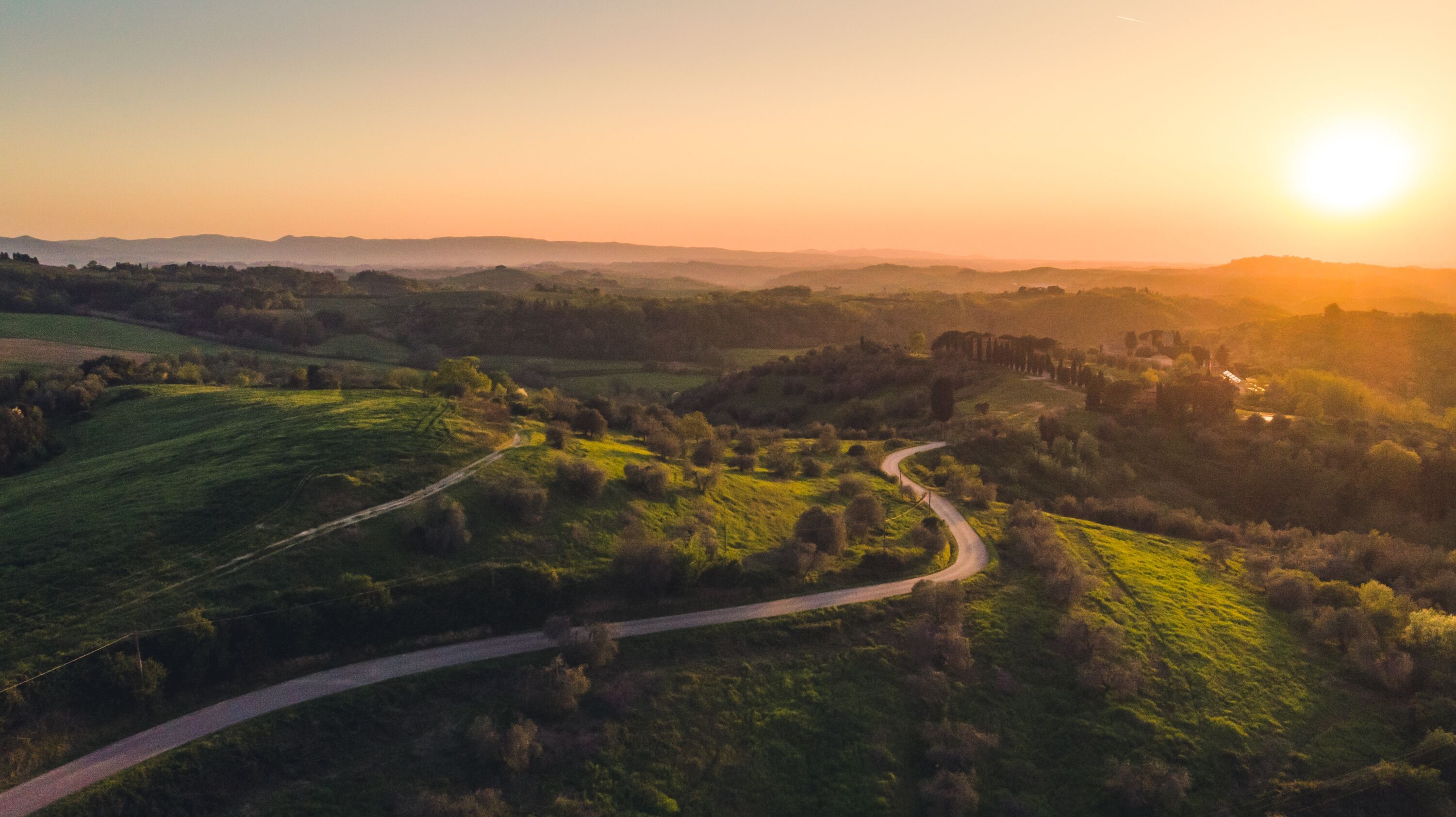 Una strada sinuosa che attraversa un paesaggio verde, con delle colline all'orizzonte e il sole al tramonto.