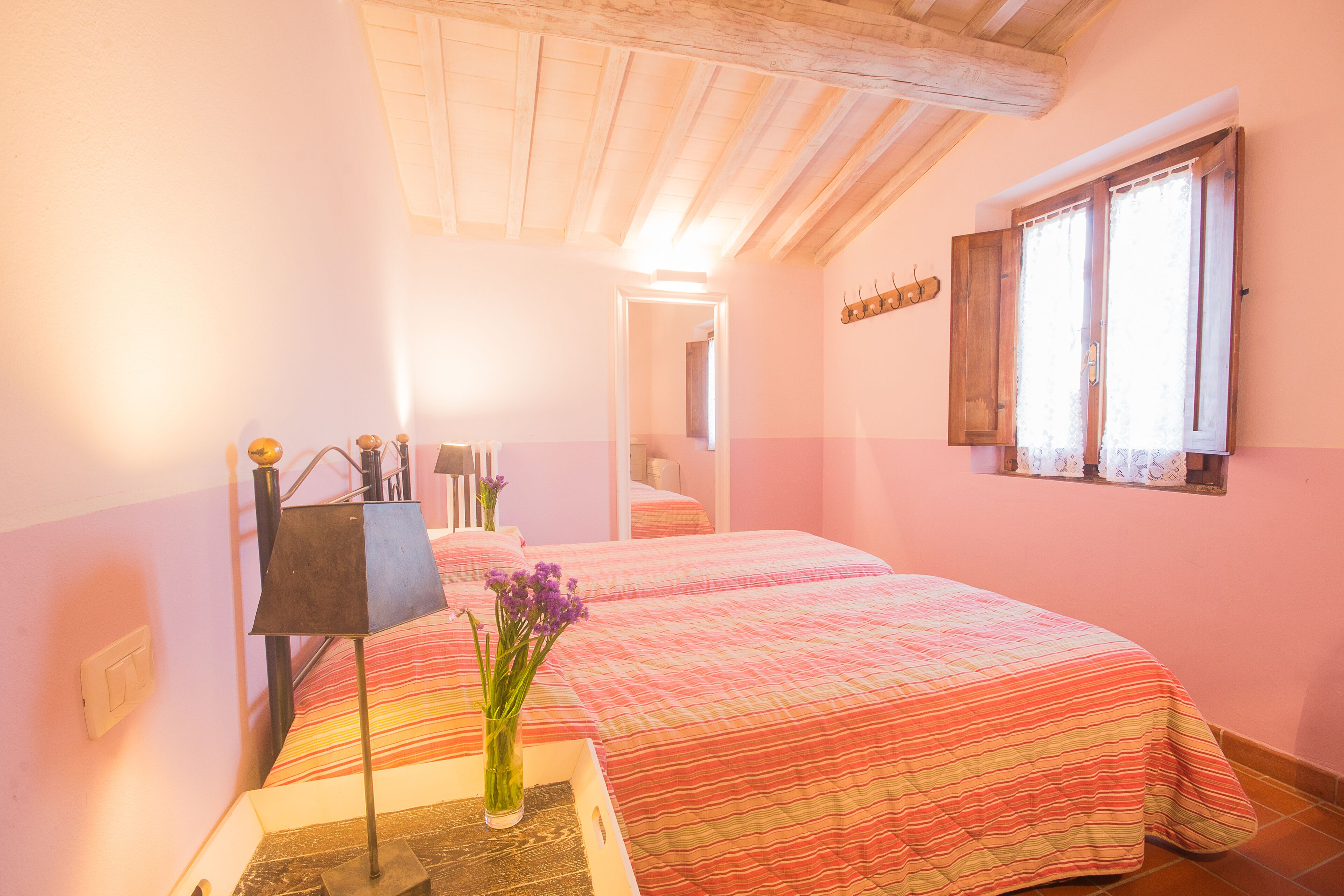 Ein Schlafzimmer mit zwei Betten, einem Nachttisch mit Lampe, einer Vase mit Blumen und einem Fenster mit Vorhängen.