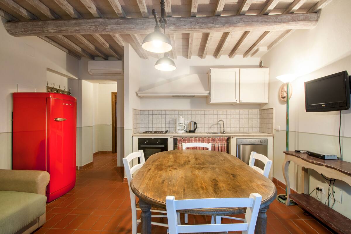 Une cuisine avec une table en bois, des chaises blanches, un réfrigérateur rouge et des placards blancs.