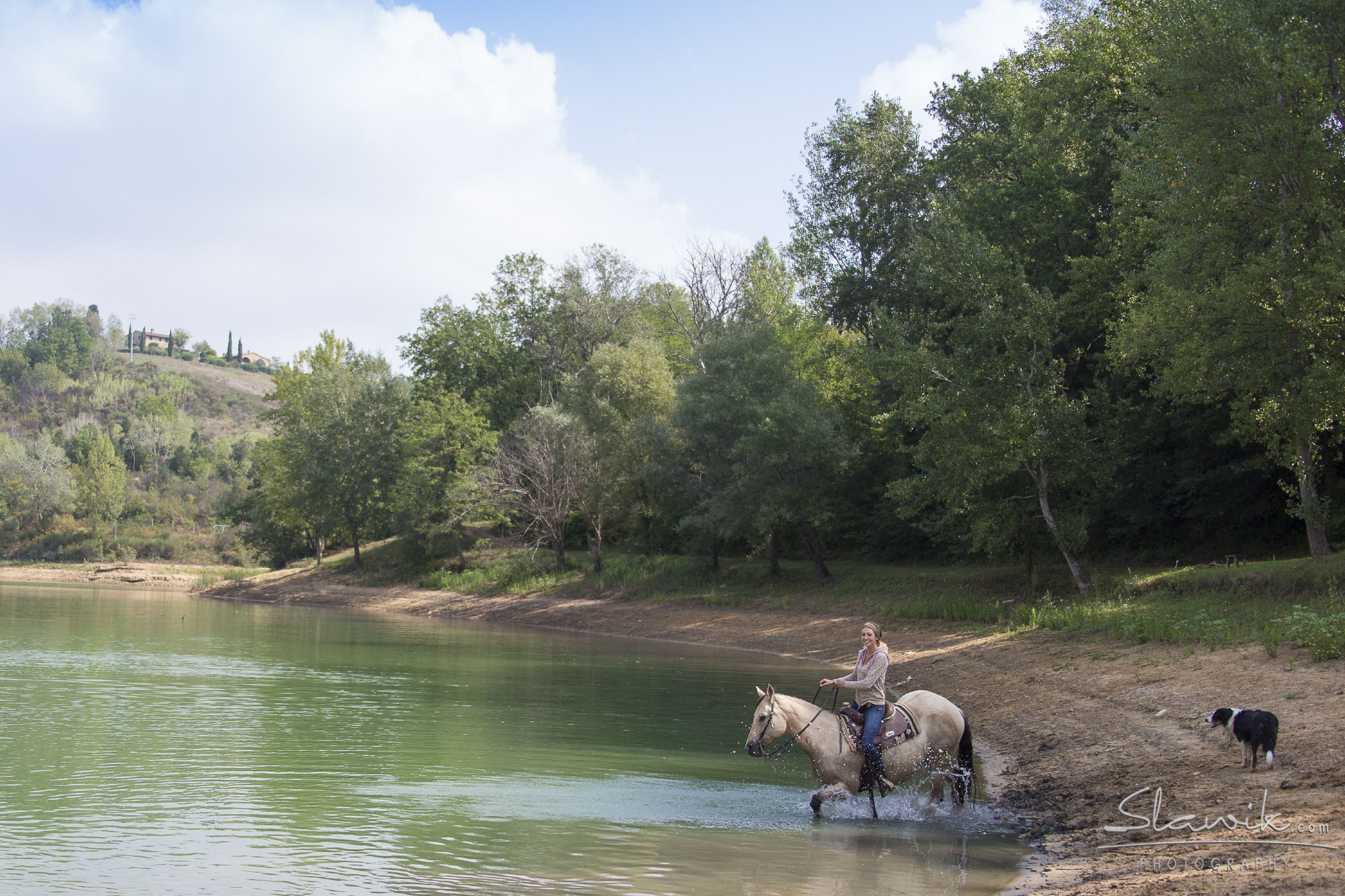 A person riding a horse through a shallow body of water, surrounded by trees and a hilly landscape.