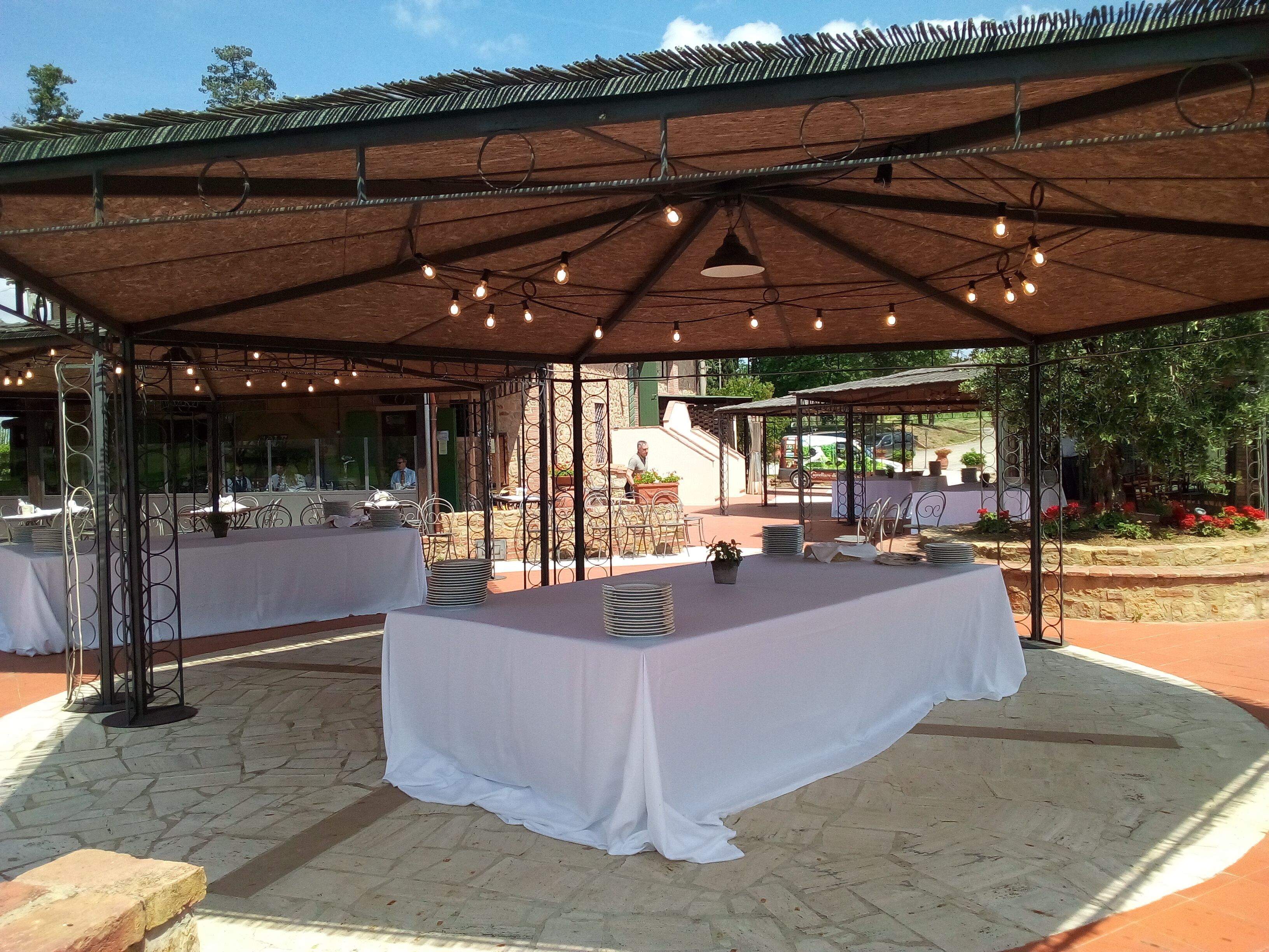 Un espacio de comedor al aire libre con mesas cubiertas con manteles blancos, bajo una estructura sombreada con luces colgantes.