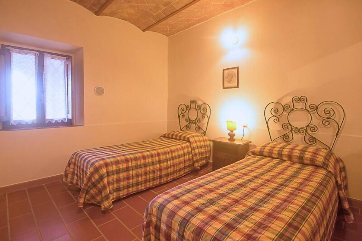 Une chambre avec deux lits, chacun doté d’une tête de lit en métal, une fenêtre ornée de rideaux en dentelle, un tableau accroché au mur et une petite table de chevet avec une lampe.