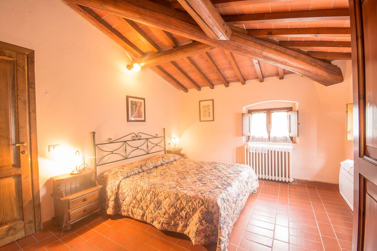 Une chambre avec un plafond en bois, un lit recouvert d’une courtepointe, une table de chevet, une fenêtre avec des rideaux et un radiateur.