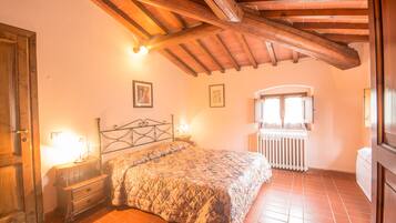 Een slaapkamer met een houten plafond, een bed met een sprei, een nachtkastje, een raam met gordijnen en een radiator.