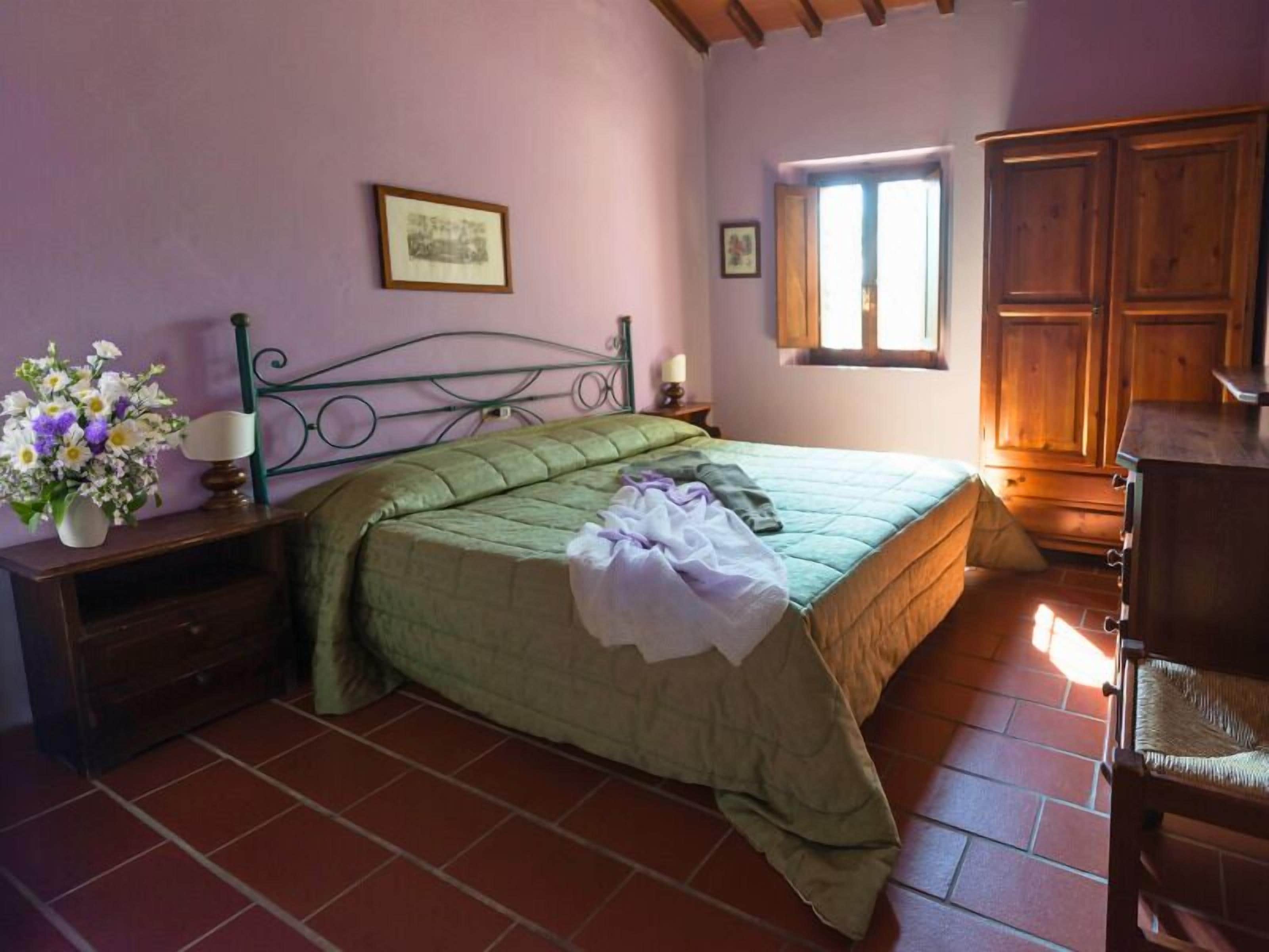 Una camera da letto con una coperta verde, un armadio in legno, una finestra con luce naturale e un comodino con fiori.