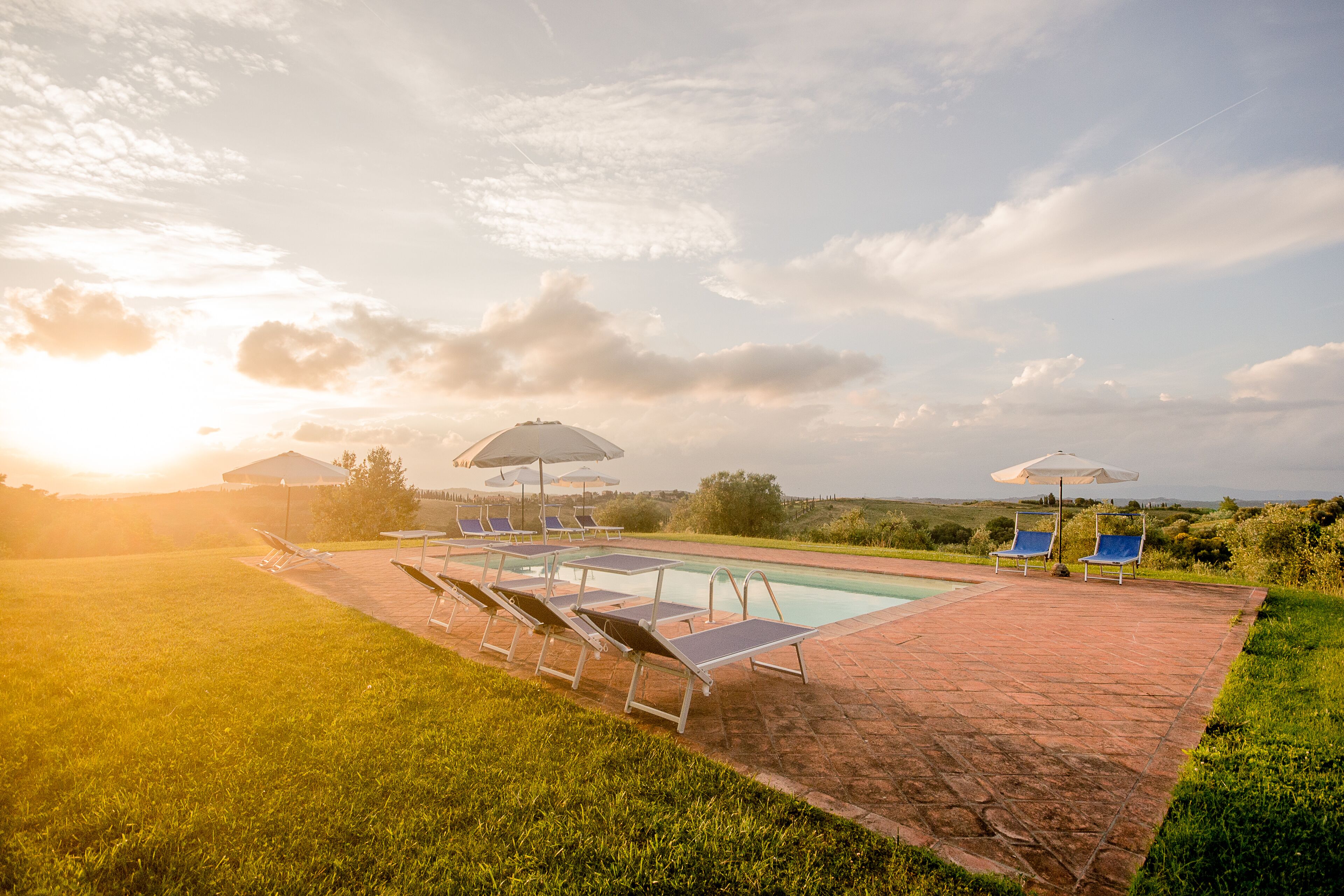 A poolside area with lounge chairs and umbrellas, surrounded by a brick patio and grassy lawn.
