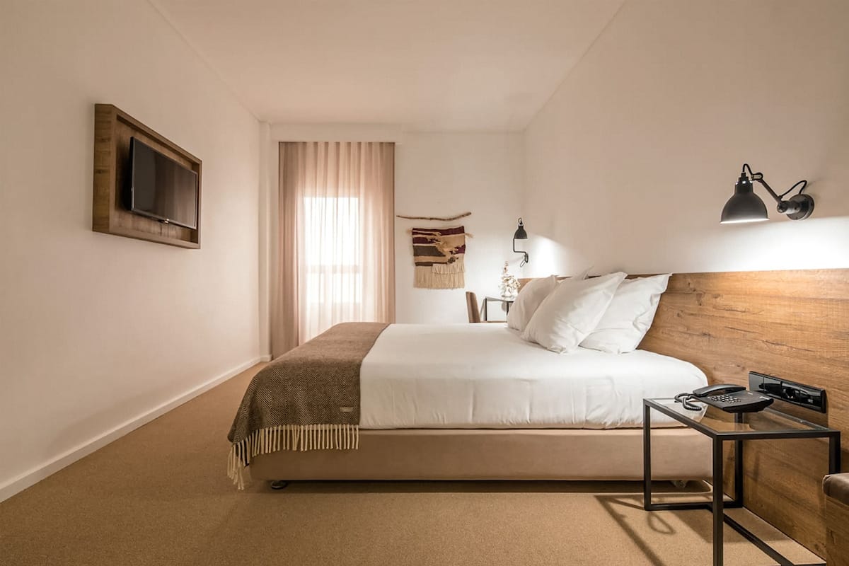 A hotel room with a bed, a bedside table with a lamp, a TV mounted on the wall, and a window with curtains.
