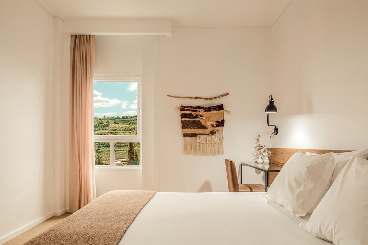 A bedroom with a large bed, a window with a view of a green landscape, a wall-mounted lamp, and a small table with a plant.