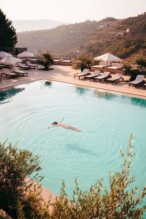 Ein Schwimmbad mit Liegestühlen und Sonnenschirmen, umgeben von viel Grün und Hügeln im Hintergrund.