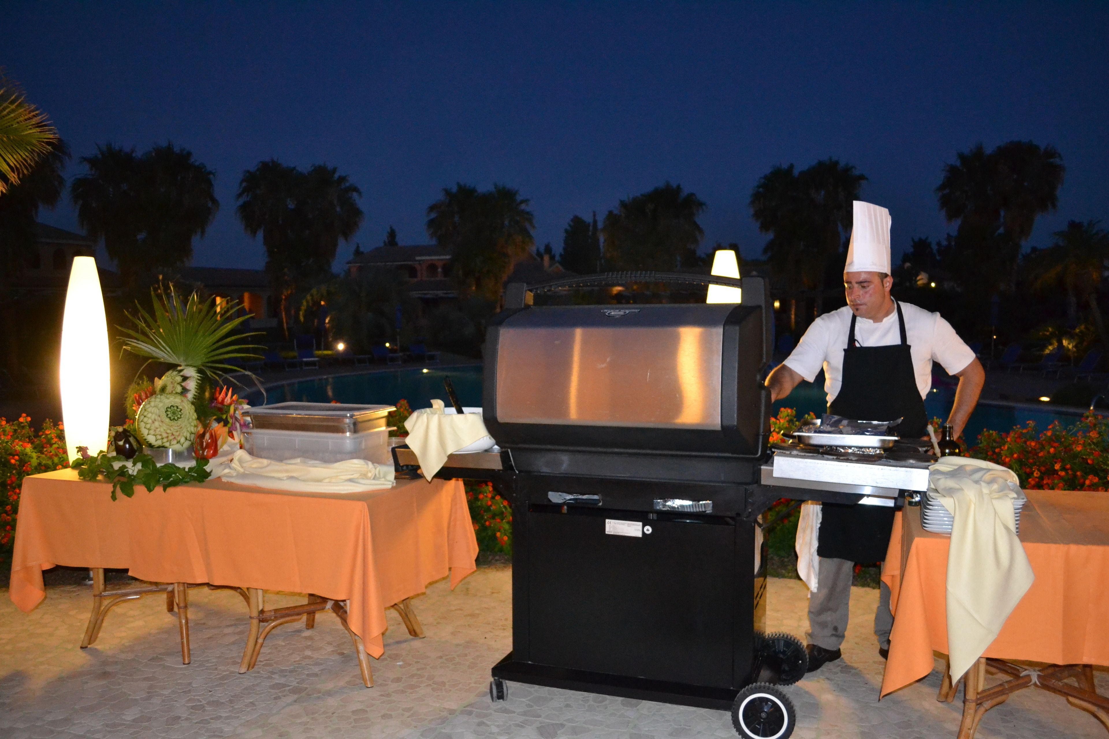 Un chef prépare des plats en plein air, la nuit, à l’aide d’un barbecue et de tables disposées pour un événement.