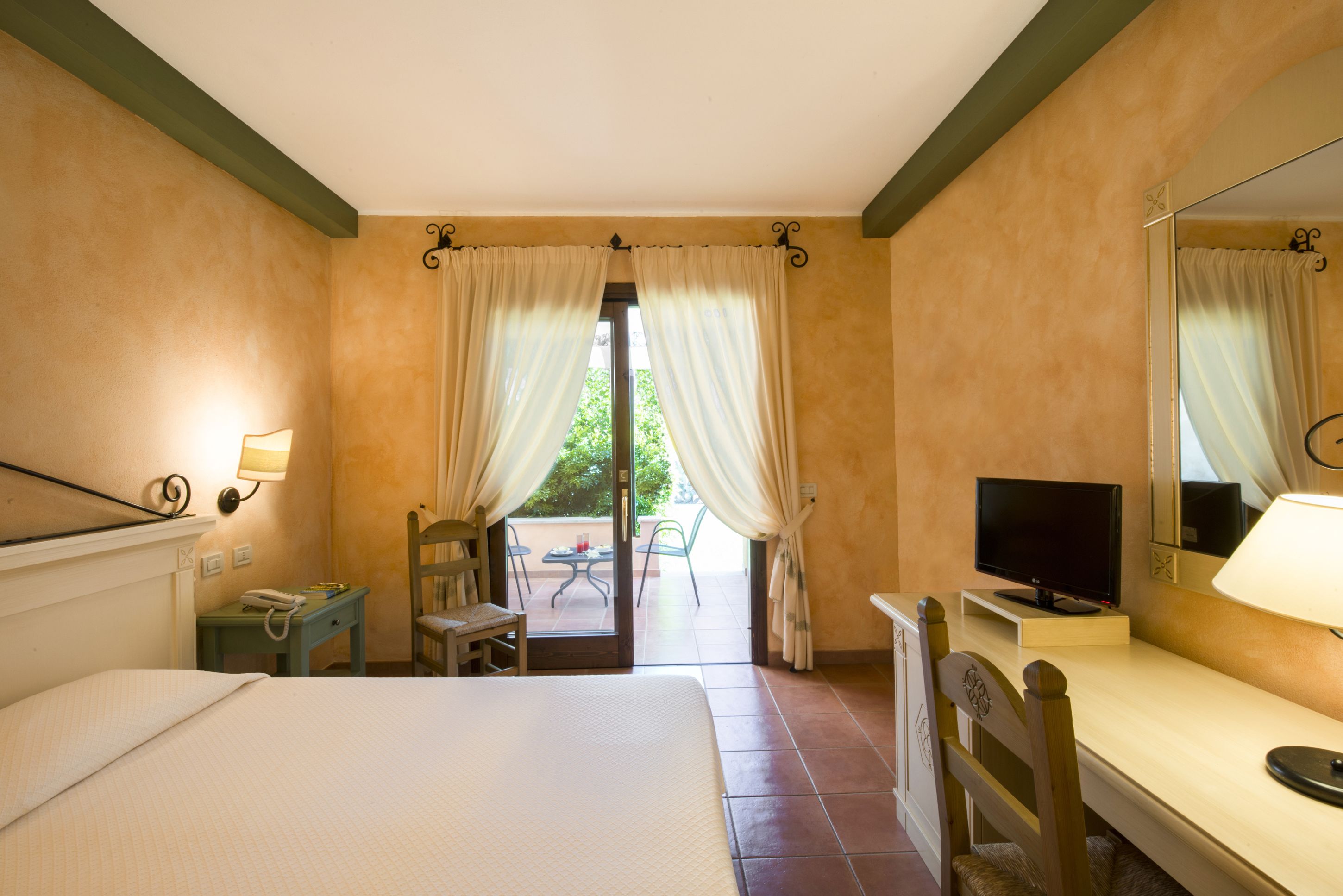 Habitación de hotel con cama, escritorio con televisor, una silla, una mesita con teléfono y ventana con cortinas.