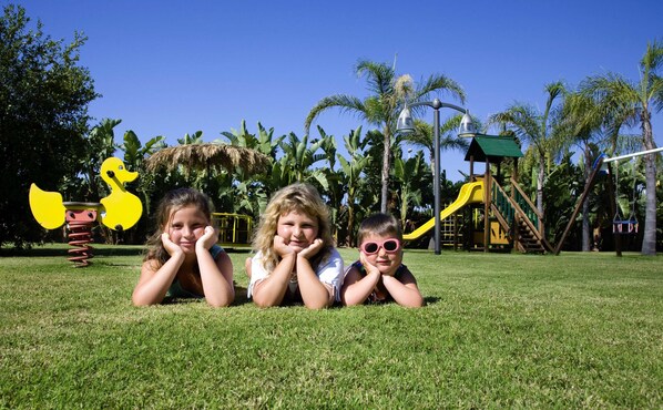 Three children lying on grass in a park with playground equipment and palm trees in the background. - Lantana Resort Hotel & Apartments (Pula)